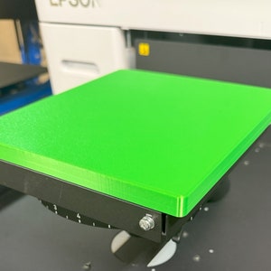 May include: A bright green square platform for a 3D printer. The platform is attached to a black base with a knob for adjusting the height.