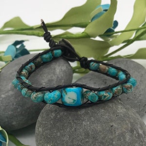 May include: A turquoise and brown beaded bracelet with a black leather cord. The bracelet is on a gray stone background.