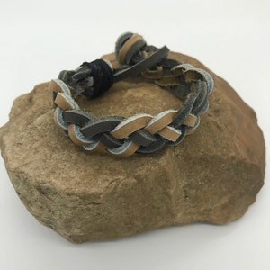 May include: A braided leather bracelet in brown and grey colours. The bracelet is tied with a knot and is displayed on a rock.