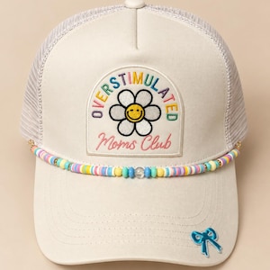 May include: A cream-colored trucker hat featuring a mesh back and an embroidered patch with a white flower, a yellow smiley face, and the text "OVERSTIMULATED Moms Club" in colorful letters. A string of multicolored beads accents the brim, with a small blue bow.