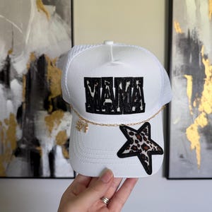 May include: White trucker cap featuring the word "MAMA" in black block letters. A gold chain with a charm hangs from the brim. A leopard print star patch is on the side.