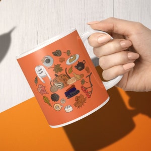 Könnte beinhalten: Orangefarbene Keramik-Tasse mit weißem Henkel, verziert mit einem herbstlichen Design. Das Design umfasst Illustrationen von Blättern, Kürbissen, einem Pullover und dem Text "London Autumn". Die Tasse wird von einer Hand gehalten.