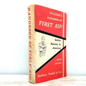 Puede incluir: Libro antiguo "Bailliere's Handbook of First Aid". La portada es roja y blanca con detalles en negro. El título está en letras blancas grandes. El libro presenta una ilustración de una persona con una lesión en el brazo. Es la quinta edición.