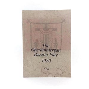 Puede incluir: Una tarjeta rectangular de papel envejecido con el texto "The Oberammergau Passion Play 1980" en negro. Un emblema rojo descolorido de una cruz y un escudo es visible en la parte superior. La tarjeta tiene un aspecto texturizado y moteado.