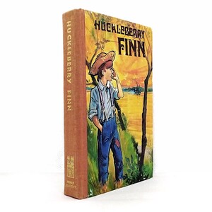 Op de afbeelding: Een hardcover boek getiteld Huckleberry Finn met een beige rug en een omslagillustratie van een persoon met een strooien hoed, lichtblauw overhemd en blauwe tuinbroek die bij zonsondergang aan een rivier staat. De titel is in zwart gedrukt.