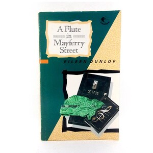 Op de afbeelding: Boekomslag van "A Flute in Mayferry Street" door Eileen Dunlop. Het ontwerp bevat een groene achtergrond, een witte titelbox en illustraties van een fluit, bladmuziek en een deur.