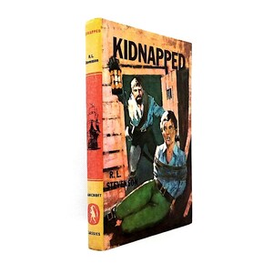 May include: A hardcover book titled Kidnapped by R.L. Stevenson with a yellow spine featuring a black ship illustration and a red circular logo. The cover art depicts a person in a blue shirt and green pants tied up while another person holds a lantern.