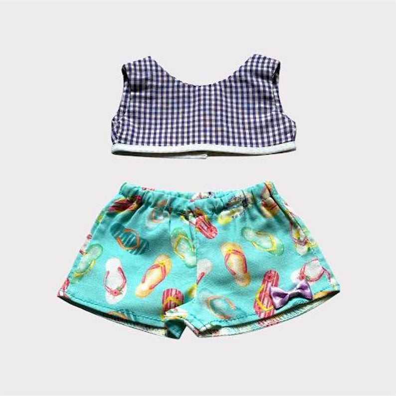 May include: A two-piece doll outfit featuring a gingham top and turquoise shorts. The shorts are patterned with colorful flip-flops and a small purple bow. The top is a navy and white checkered pattern.