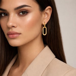May include: Gold-colored oval hoop earrings. The earrings are connected by a small, round hoop at the top. The oval shape has a textured surface. The earrings are worn by a person wearing a tan blazer.