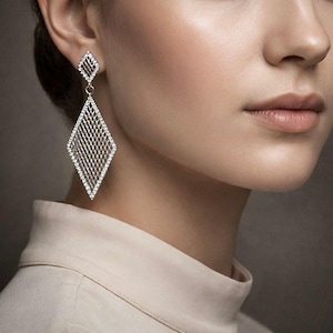 May include: A diamond-shaped earring with a delicate, open-weave design. The earring is adorned with small, sparkling stones, creating a shimmering effect. The earring hangs from a smaller, diamond-shaped stud. The background is a soft, neutral tone.