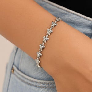 Puede incluir: Una pulsera plateada con una serie de pequeños dijes de tortugas. La pulsera se lleva en la muñeca, con las tortugas unidas entre sí. La pulsera está hecha de un material metálico brillante.