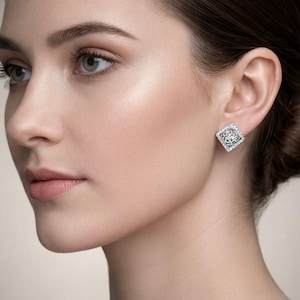 May include: A close-up of a person's face with a square-shaped earring. The earring is silver-toned with a detailed, openwork design in the center, surrounded by small, sparkling stones. The earring is set against the person's ear.