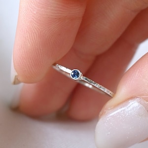 Könnte beinhalten: Ein silberner Ring mit einem kleinen blauen Edelstein in der Mitte. Der Ring ist dünn und zart, mit einer strukturierten Oberfläche.