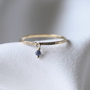 Könnte beinhalten: Ein zarter Goldring mit einem kleinen blauen Edelstein, der daran hängt. Der Ring liegt auf einem weißen Stoffhintergrund.