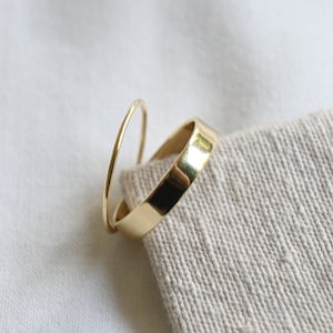 Könnte beinhalten: Zwei goldene Ringe, einer dünn und einer dick, liegen übereinander. Die Ringe liegen auf einer weißen Stoffoberfläche.