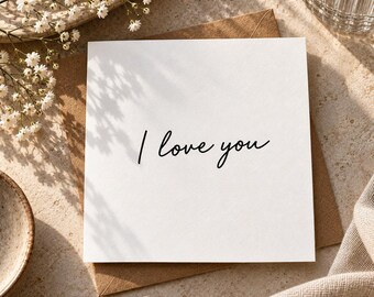 May include: A square white greeting card featuring the black cursive text I love you, positioned on top of a brown kraft paper envelope. The card has a clean, minimalist design with a smooth texture, presented as a simple folded paper stationery item.