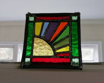 Könnte beinhalten: Ein farbenfrohes Buntglasfenster mit einem Sonnenaufgangsmotiv. Das Design zeigt einen Halbkreis aus gelbem Glas am unteren Rand, mit Strahlen in Orange, Rot, Lila, Blau und Grün. Das Panel ist schwarz umrandet und hat eine Kette zum Aufhängen.