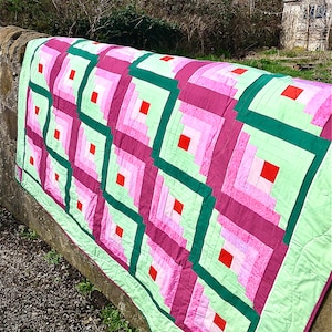 May include: A quilt with a log cabin pattern in shades of pink, green, and red. The quilt is made of fabric squares sewn together to create a geometric design.
