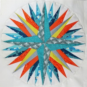 May include: A teal, orange, yellow, and gray starburst quilt block with a white background. The block is made of fabric with a variety of patterns, including floral, geometric, and paisley.