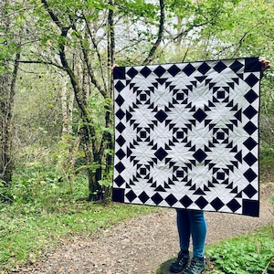 May include: A monochrome quilt featuring a geometric design of black diamonds and triangles on a white background. The quilt is displayed outdoors, held up against a backdrop of trees. The quilt measures approximately 152 cm by 152 cm.