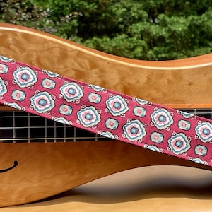 Puede incluir: Una correa de guitarra roja con un patrón geométrico blanco y azul. La correa está sobre una guitarra de madera.