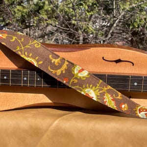 Puede incluir: Una correa de guitarra con estampado floral marrón y crema. La correa está sobre un instrumento musical de madera con fondo marrón.