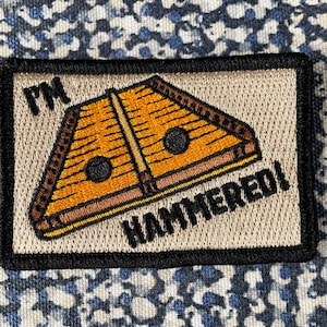 May include: Embroidered patch featuring a hammered dulcimer design in shades of orange and brown, with the text "FN HAMMERED!" in black. The patch has a black border and a tan background.