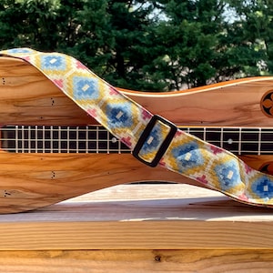 Puede incluir: Un dulcémele de madera con una correa estampada con diseños geométricos azules, amarillos y rosas. La correa tiene una hebilla negra. El dulcémele tiene un acabado de madera natural e incrustaciones florales decorativas.
