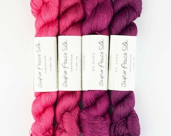 Peut inclure: Quatre écheveaux de fil Jaipur Peace Silk disposés côte à côte dans un dégradé allant du rose vif au violet foncé. Chaque écheveau porte une étiquette en papier blanc avec le texte noir Jaipur Peace Silk, BC Garn et ca. 300m / 50g.
