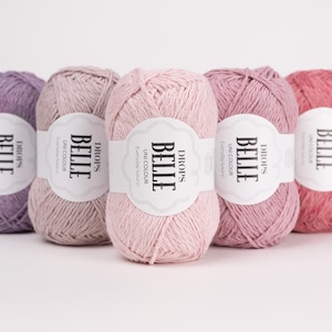 May include: Five balls of Drops Belle yarn in shades of purple, pink and coral. Each ball has a white label with the text "BELLE" and "DROPS". The yarn is for knitting and crochet projects. The yarn is in shades of lavender, light pink, medium pink, mauve, and coral.