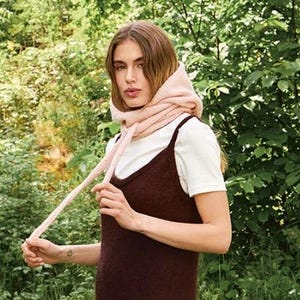 Op de afbeelding: Een vrouw draagt een donkerbruine maxi-jurk met een hoge zijsplit, over een wit t-shirt. Ze draagt ook een lichtroze sjaal en bruine schoenen. De achtergrond is een weelderig groen bos.