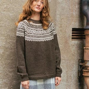 May include: A woman wearing a brown sweater with a white geometric pattern around the neckline, a gray plaid skirt, dark gray tights, and red shoes. The sweater is long-sleeved and appears to be made of a knit material. The background is a textured wall.