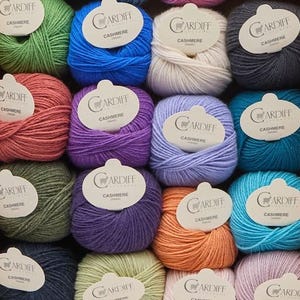 May include: A cardboard box containing twenty-five individual balls of yarn in various colors. Each ball features a white paper label with the text Cardiff Cashmere printed on it. The yarn balls are arranged in a grid pattern within the box.
