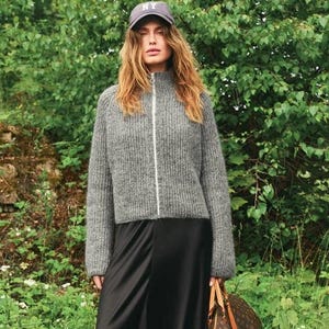 May include: A woman wearing a gray zip-up sweater, black skirt, and a baseball cap with "NY" on it, standing in a grassy area. She is holding a brown and tan patterned tote bag with tan straps.