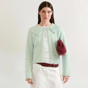 May include: A woman wearing a light green cardigan with a scalloped collar, a white top, and a white skirt. She is holding a small, burgundy purse. A brown belt and tan flats complete the outfit. A stack of books is on the floor.