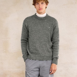 Può includere: Un uomo indossa un maglione grigio, pantaloni grigi e scarpe da ginnastica bianche e verde neon. È in piedi davanti a un muro color beige chiaro.