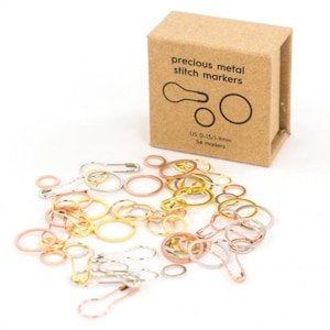 Pode incluir: Um conjunto de 54 marcadores de ponto de metal precioso em ouro, prata e ouro rosa. Os marcadores vêm em uma variedade de tamanhos, incluindo os tamanhos americanos 0-15/1-9 mm. Os marcadores são embalados em uma pequena caixa de papelão com o texto "precious metal stitch markers" impresso na frente.