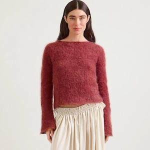 Può includere: Una donna indossa un maglione bordeaux a maniche lunghe e pantaloni a gamba larga color crema. Il maglione ha uno scollo tondo e una consistenza soffice. I pantaloni sono a vita alta e fluidi, creando una silhouette rilassata. L'outfit è semplice ed elegante.