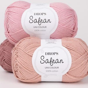 May include: Three balls of Drops Safran yarn in varying shades of pink and peach. Each ball has a white label with the text "DROPS Safran UNI COLOUR 100% cotton". The yarn is tightly wound, showcasing the texture of the cotton fibres.