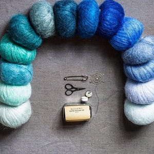 May include: An arch of fuzzy yarn balls in a gradient of blue and teal shades, ranging from deep navy to pale white. Below the arch are a silver safety pin, black scissors, a small metal snowflake ornament, and a rectangular tin labelled Rapid Repair Kit.