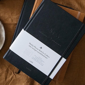 May include: Two skeins of dark green yarn labeled "Novita Laine" and a black planner with the text "2023 Weekly Planner Built for Crafters" are displayed on a brown fabric surface. A small white plate holds craft supplies.