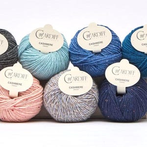 May include: Nine balls of Cardiff Cashmere yarn arranged in two rows. Each ball features a white paper label with the text Cardiff Cashmere. The yarn colors include charcoal, light blue, medium blue, dark blue, pink, grey, and white with speckles.