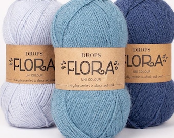 May include: Three skeins of yarn in varying shades of blue, including light blue, medium blue, and dark blue. Each skein features a tan paper label with the text DROPS FLORA UNI COLOUR and the phrase Everyday comfort in alpaca and wool.
