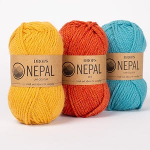 Może przedstawiać: Trzy motki przędzy Drops Nepal w kolorze żółtym, pomarańczowym i turkusowym. Każdy motek ma brązową etykietę z napisem "DROPS NEPAL" i "Wool and alpaca for everyday". Przędza to mieszanka kolorów uni.