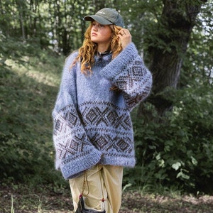 May include: A blue and brown patterned sweater with a high neck and long sleeves. The sweater has a relaxed fit and is made of a soft, fuzzy material. The model is wearing a black baseball cap and beige pants.