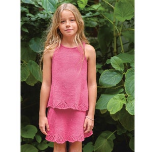 May include: A young girl wearing a pink sleeveless top and skirt with a scalloped hem. The top and skirt are both knitted and have a delicate texture. The girl is standing in front of a green leafy background.