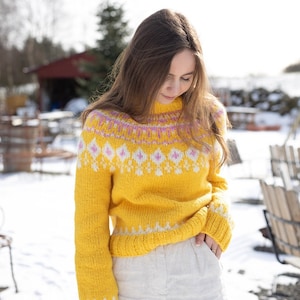May include: A bright yellow long-sleeve knit sweater featuring a high neckline and a decorative yoke pattern. The design includes white and pink diamond-shaped motifs across the chest and shoulders, with matching white geometric patterns at the cuffs and hem.