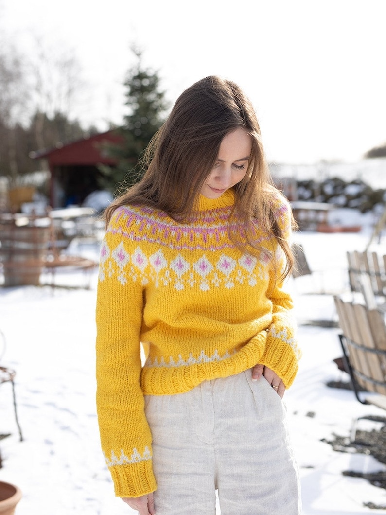 May include: A bright yellow long-sleeve knit sweater featuring a high neckline and a decorative yoke pattern. The design includes white and pink diamond-shaped motifs across the chest and shoulders, with matching white geometric patterns at the cuffs and hem.