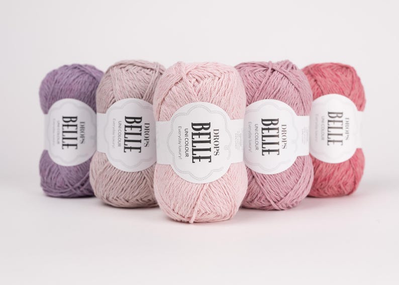 May include: Five balls of Drops Belle yarn in shades of purple, pink and coral. Each ball has a white label with the text "BELLE" and "DROPS". The yarn is for knitting and crochet projects. The yarn is in shades of lavender, light pink, medium pink, mauve, and coral.