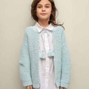May include: A light blue knitted cardigan with long sleeves and a tie detail at the neckline. The cardigan has tassels on the tie and is worn over a white button-down shirt.